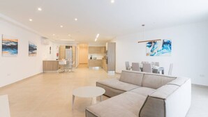 A spacious living area with a sofa, a round coffee table, and a dining table set for four. There is a kitchen area in the background with modern appliances and cabinets. - Property With Pool Access - Xaghra Near Marsalforn (Xaghra)