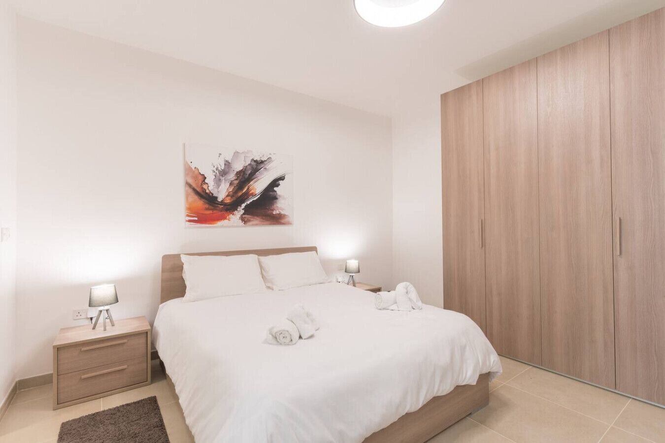A modern bedroom with a large bed, a nightstand with a lamp, a wooden wardrobe, and a painting on the wall.