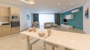 A modern kitchen with built-in appliances, a dining area with a table and chairs, a living room with a sofa and a TV, and a teal accent wall. - Property With Pool Access - Xaghra Near Marsalforn (Xaghra)