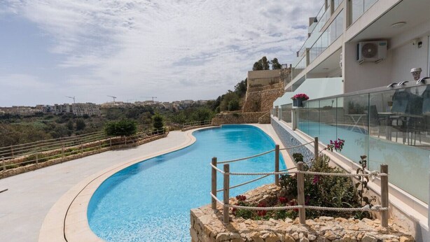 A curved swimming pool with a clear view of the surrounding landscape, a stone wall, and a modern building with glass railings. - Property With Pool Access - Xaghra Near Marsalforn (Xaghra)