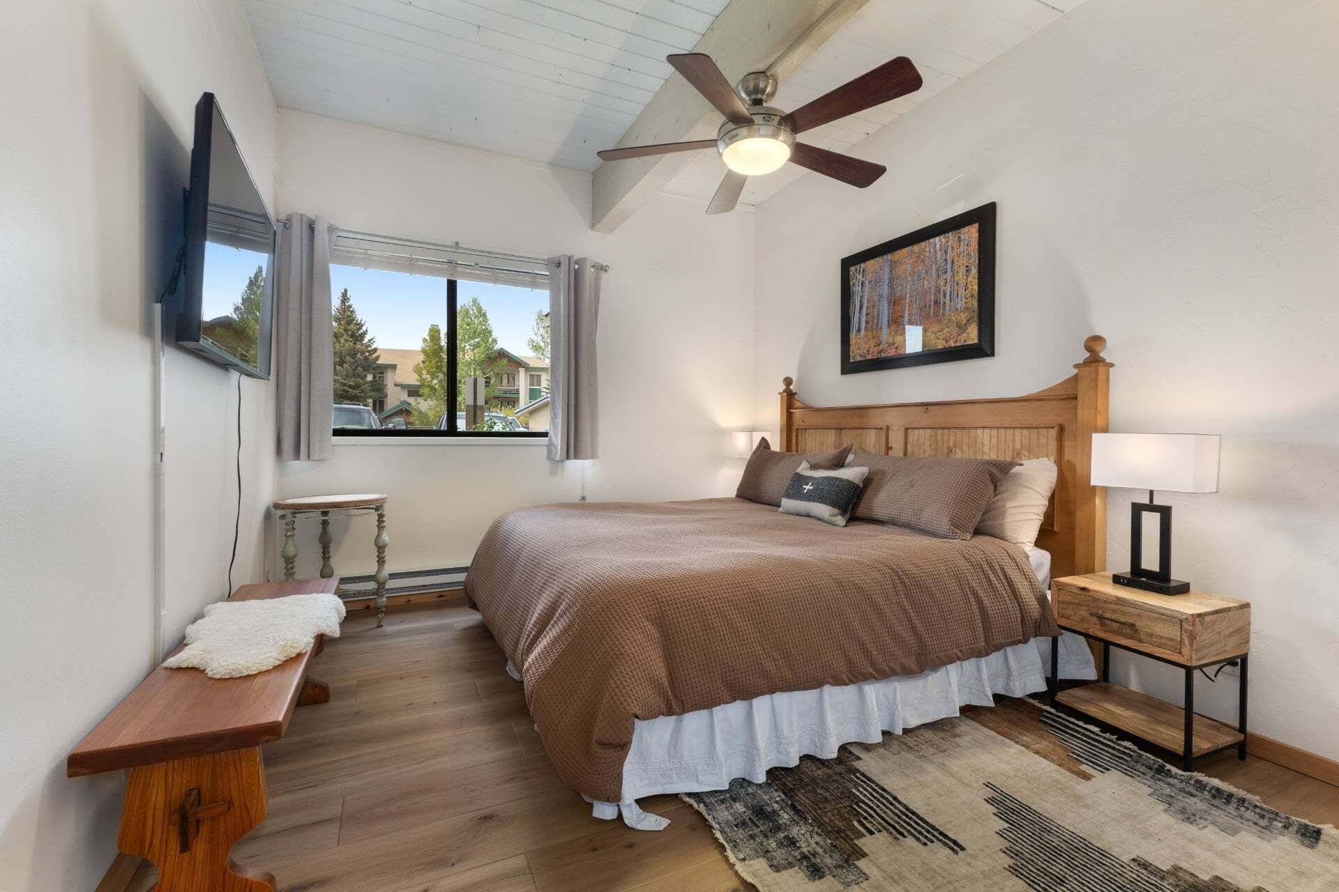Quarto com cama, mesas de cabeceira, ventilador de teto, janela com cortinas, um quadro e vista para o exterior.