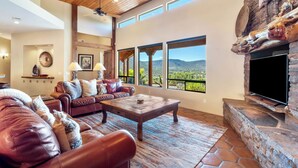 Un salon spacieux doté d’une cheminée en pierre, de canapés en cuir et de grandes fenêtres offrant une vue sur les montagnes.