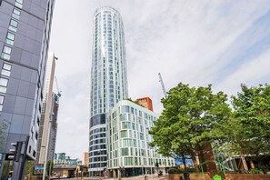 A modern high-rise building with a curved glass facade, surrounded by other buildings and trees. - Blueground | Lambeth, rooftop, nr Vauxhall (London)