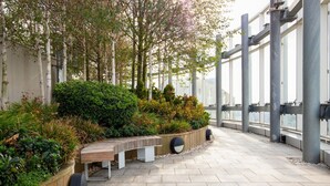 Une terrasse extérieure avec un banc en bois, des jardinières contenant diverses plantes et une vue sur la ville à travers de grandes fenêtres.