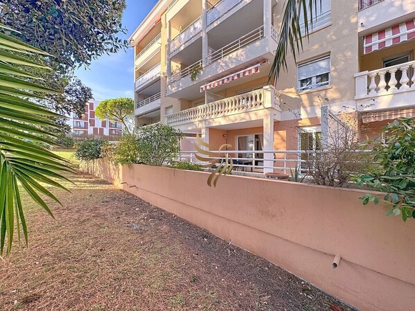 A multi-story building with balconies, a garden area with a low wall, and a clear sky.
