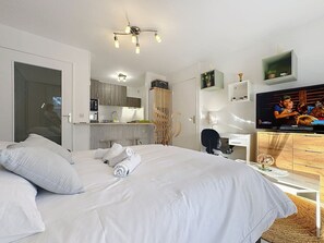 A modern hotel room with a large bed, a television, a desk with a chair, and a kitchenette in the background.