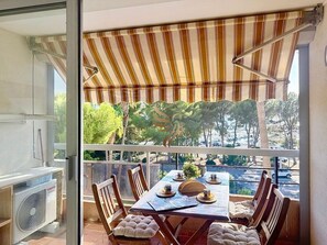 Ein Balkon mit gestreiftem Jalousievorhang, einem Esstisch gedeckt mit Tellern, Tassen und einer Vase, und Blick auf Bäume und eine Straße.