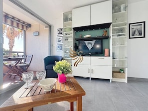Uma sala de estar com uma mesa de madeira, uma cadeira azul, um sofá e uma TV sobre um móvel.