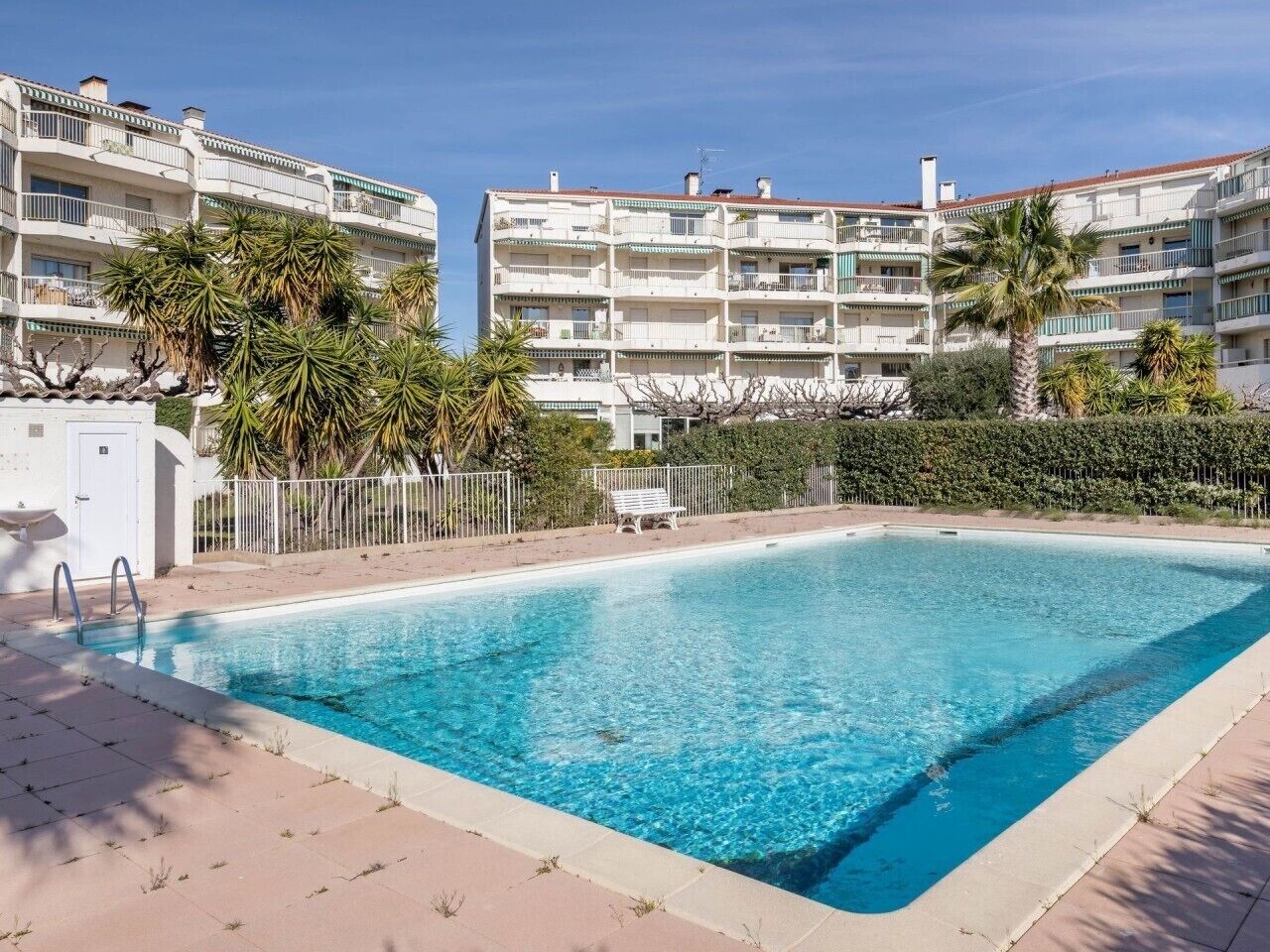 A rectangular pool with a clear blue water surface, surrounded by a tiled deck. In the background, there are multi-story buildings with balconies and a hedge.