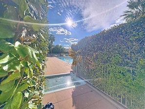 Uma área de piscina com deck revestido, cercada por vegetação densa e uma cerca de metal.