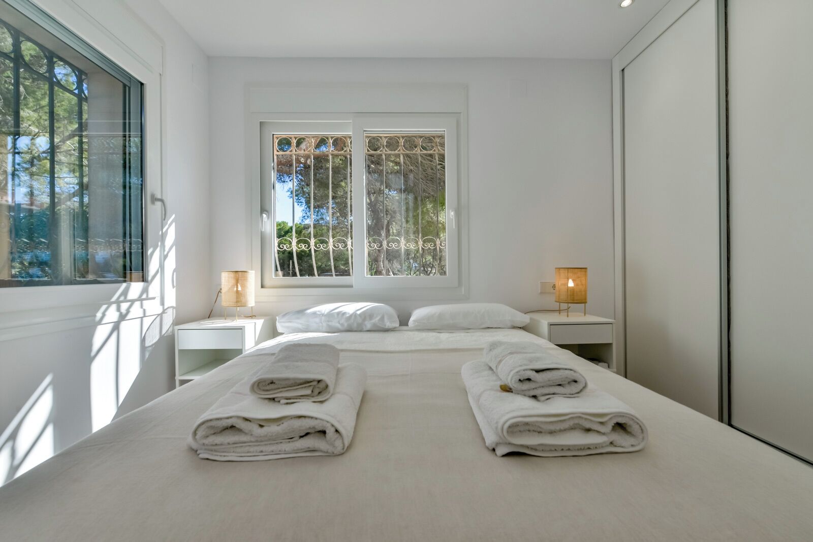 A modern bedroom with a large bed, two bedside tables, a window with decorative bars, and a view of trees.