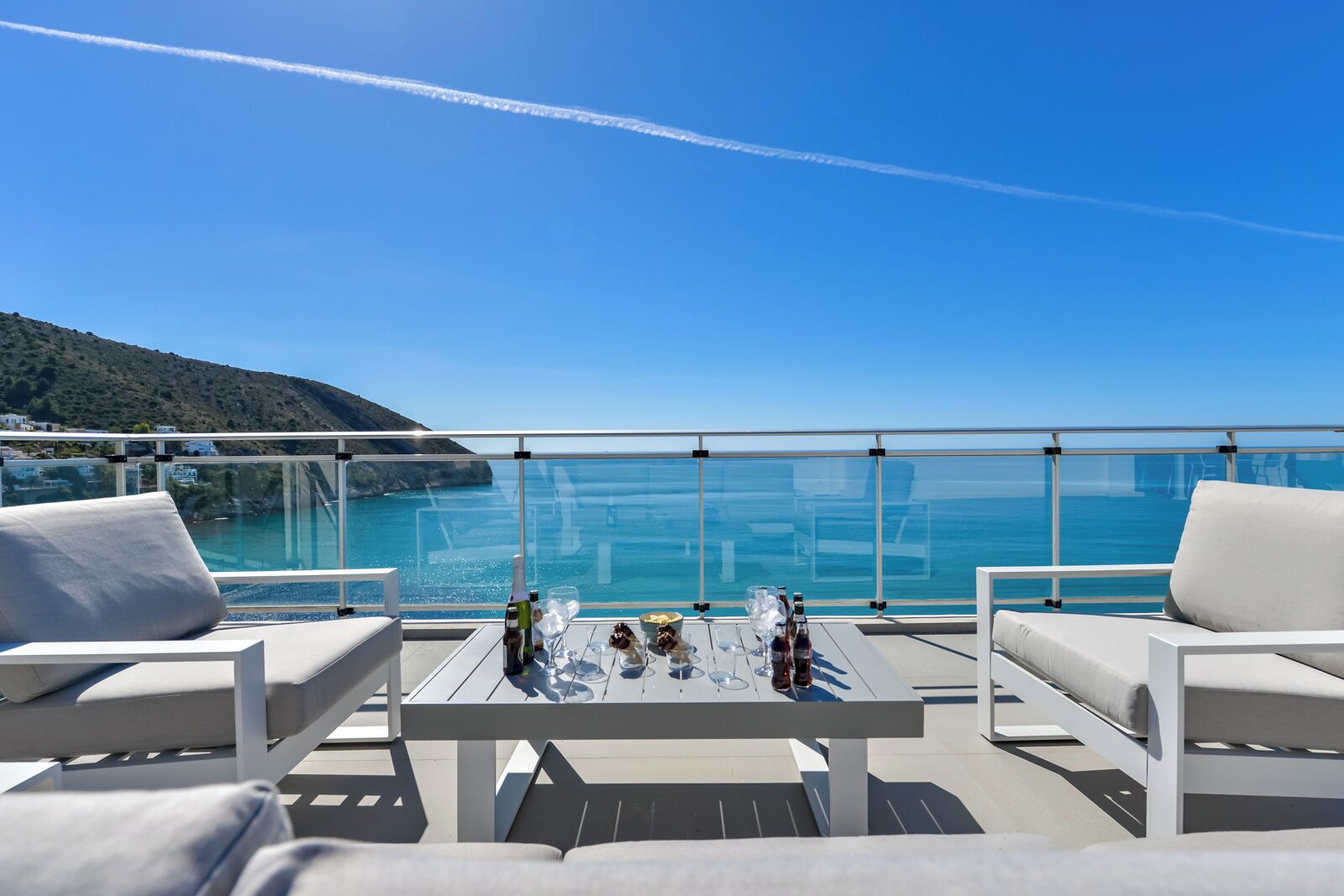 A balcony with white furniture, a table with bottles and glasses, and a clear view of the sea and a distant coastline.