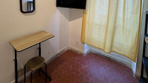 A small room with a tiled floor, a wooden desk, a chair, a wall-mounted TV, and a yellow curtain. - Nuitéa Résidence  (Marseille)