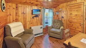 A cabin interior with wooden walls, a ceiling fan, a television, a couch, a chair, and a table. - Hedgehog (Wilson Pond Cabins) (Greenville)