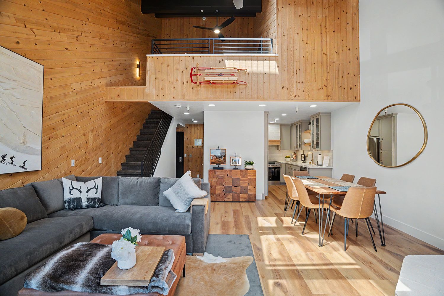 A modern interior with a staircase leading to an upper level, a dining area with a wooden table and chairs, and a living room with a sofa and coffee table.