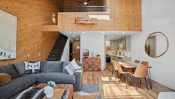 A modern interior with a staircase leading to an upper level, a dining area with a wooden table and chairs, and a living room with a sofa and coffee table.