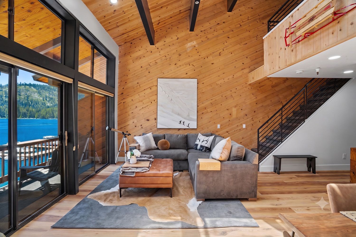 A modern living room with a large window overlooking a lake, a gray sectional sofa, a wooden coffee table, and a staircase leading to an upper level.