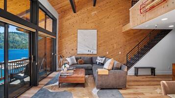A modern living room with a large window overlooking a lake, a gray sectional sofa, a wooden coffee table, and a staircase leading to an upper level.