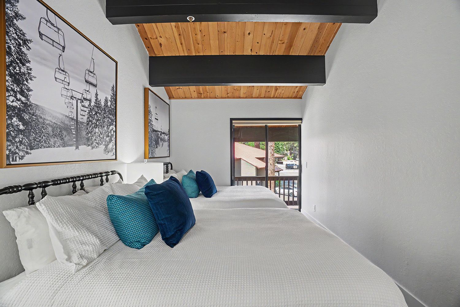 A bedroom with a large bed, wooden ceiling beams, a balcony with a railing, and a view of the outside.