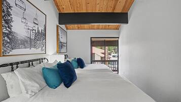 A bedroom with a large bed, wooden ceiling beams, a balcony with a railing, and a view of the outside.