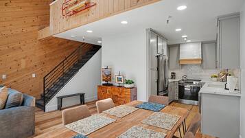 Cozinha moderna com eletrodomésticos de aço inoxidável, sala de jantar com mesa e cadeiras de madeira e escada que leva a um andar superior.