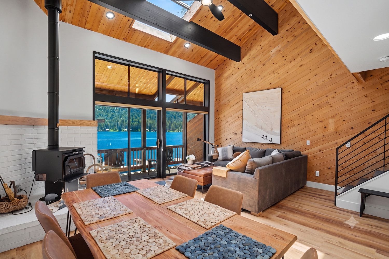 Un intérieur moderne avec un plafond en bois, une grande fenêtre donnant sur un plan d’eau, une cheminée et un coin salon comprenant un canapé et une table.