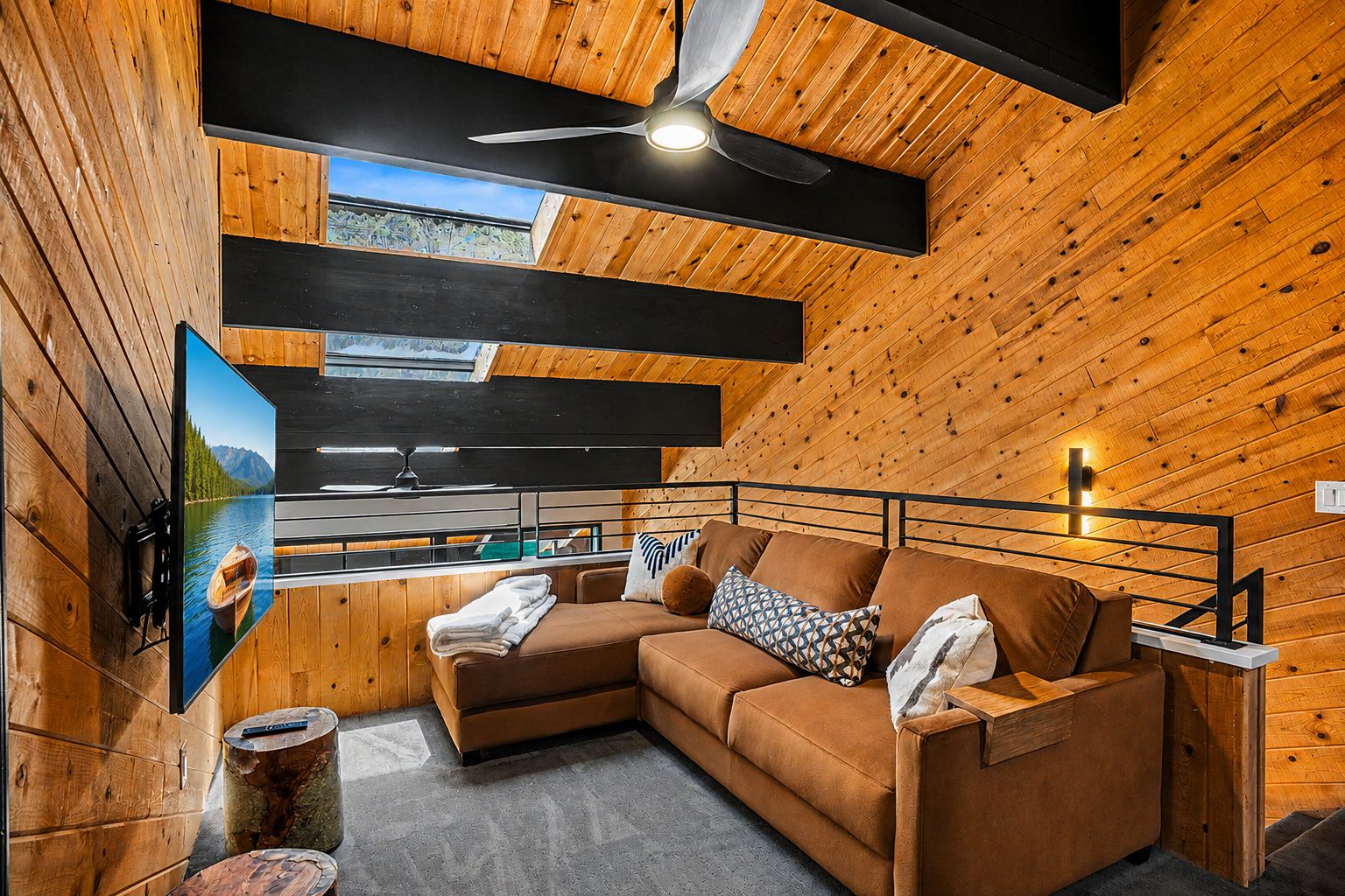 A cozy cabin interior with a wooden ceiling, a ceiling fan, a sofa with cushions, a mounted television, and a window overlooking a forested area.