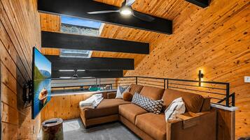 A cozy cabin interior with a wooden ceiling, a ceiling fan, a sofa with cushions, a mounted television, and a window overlooking a forested area.