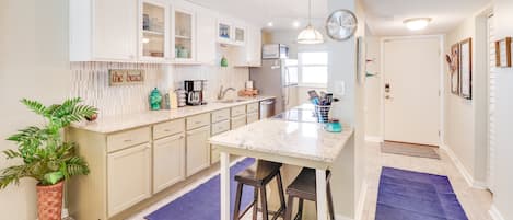 A modern kitchen with white cabinets, a granite countertop, and a central island. There is a potted plant and a blue rug on the floor.