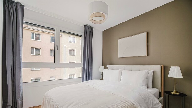 Ett hotellrum med en stor säng, ett nattduksbord med en lampa, utsikt över staden genom ett fönster och en tavla som hänger på väggen.