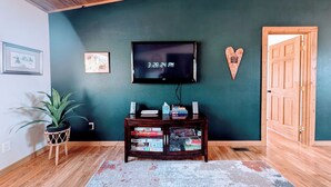 Una habitación con techo de madera, un televisor montado en la pared, una puerta de madera, una planta pequeña y una estantería con libros y juegos.