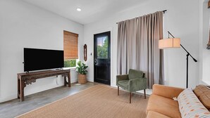 Una sala de estar con televisor de pantalla plana, una mesa de centro de madera, un sillón verde, un sofá beige, una lámpara de pie y un ventanal con cortinas.
