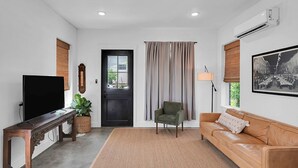 Una sala de estar con televisión, un sofá, una silla, una puerta y una ventana con cortinas.
