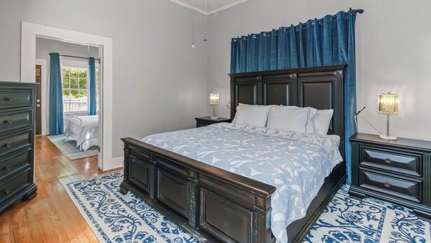 Un dormitorio con una cama grande, cabecera de madera, dos mesitas de noche con lámparas, un tapete azul con estampado y una puerta que da a otra habitación.