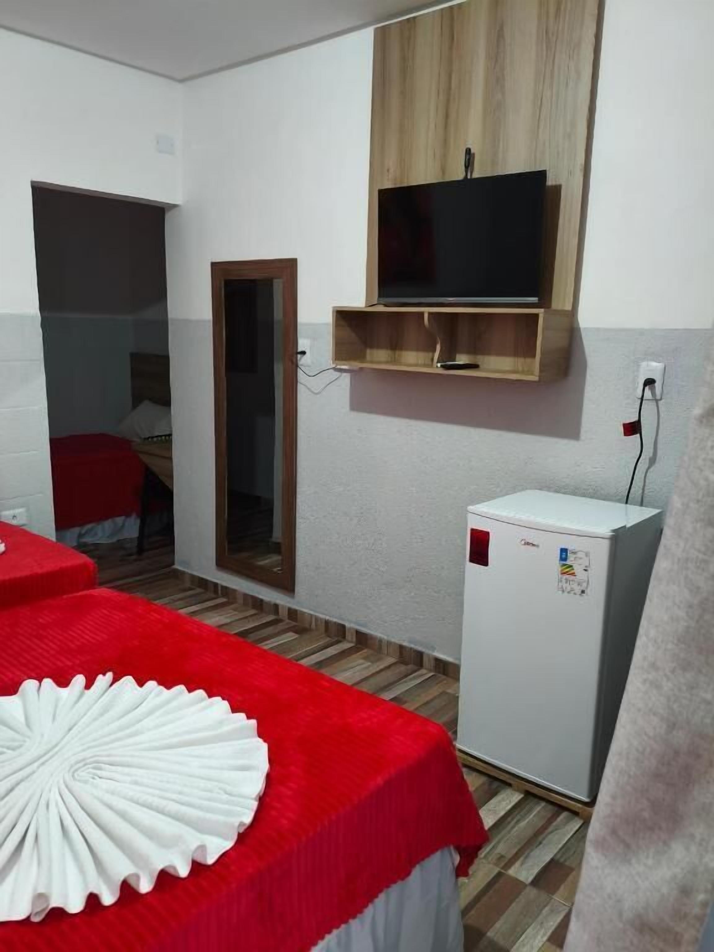 A small room with a bed, a television mounted on a wooden shelf, a refrigerator, and a mirror.
