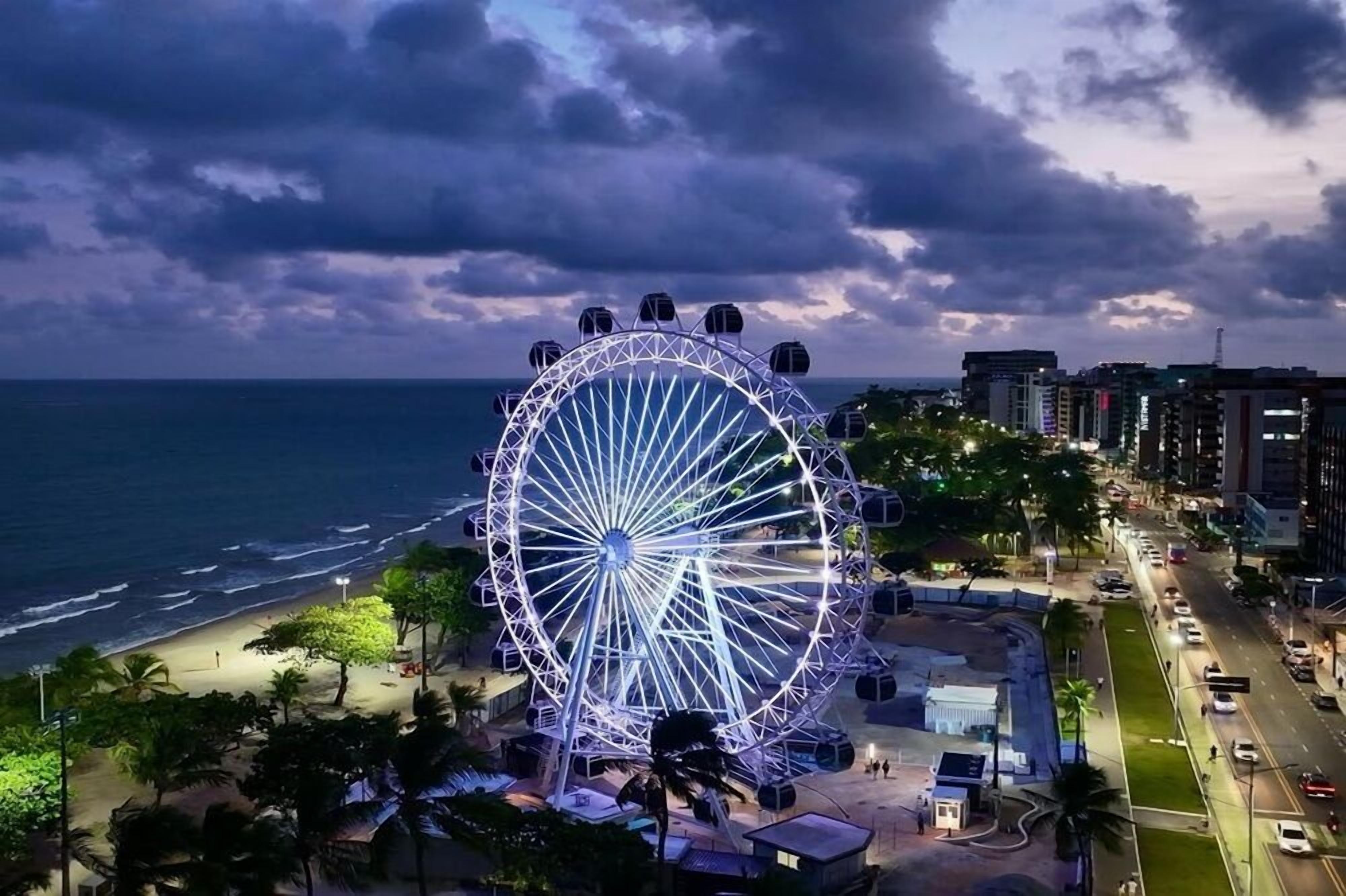 Ein großes Riesenrad mit einer kreisförmigen Struktur und mehreren Gondeln, gelegen in der Nähe eines Strandes und einer Stadt mit Gebäuden und Straßen.