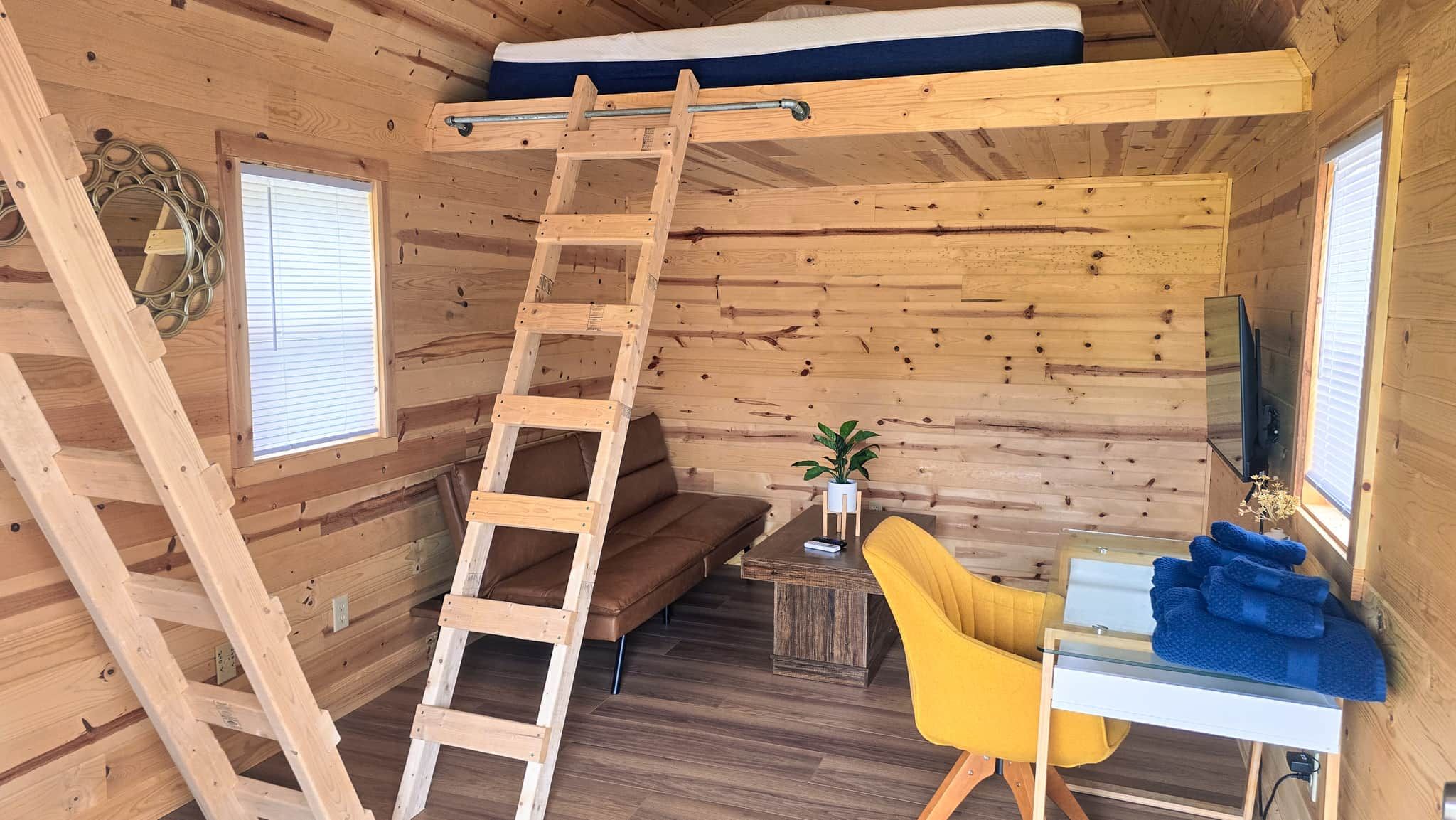 Interior de cabana com cama no mezanino, escada de madeira, sofá, mesinha com uma planta, cadeira amarela e TV na parede.