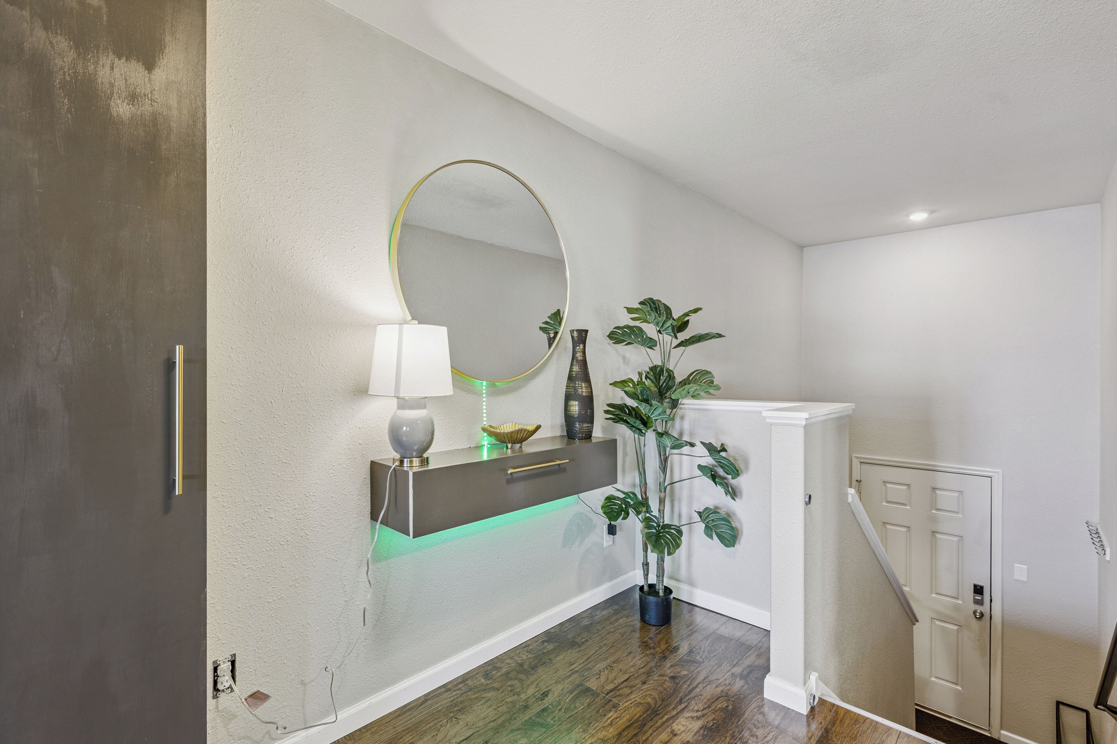 A hallway with a white wall, a wooden floor, a mirror, a lamp, a plant, and a door.