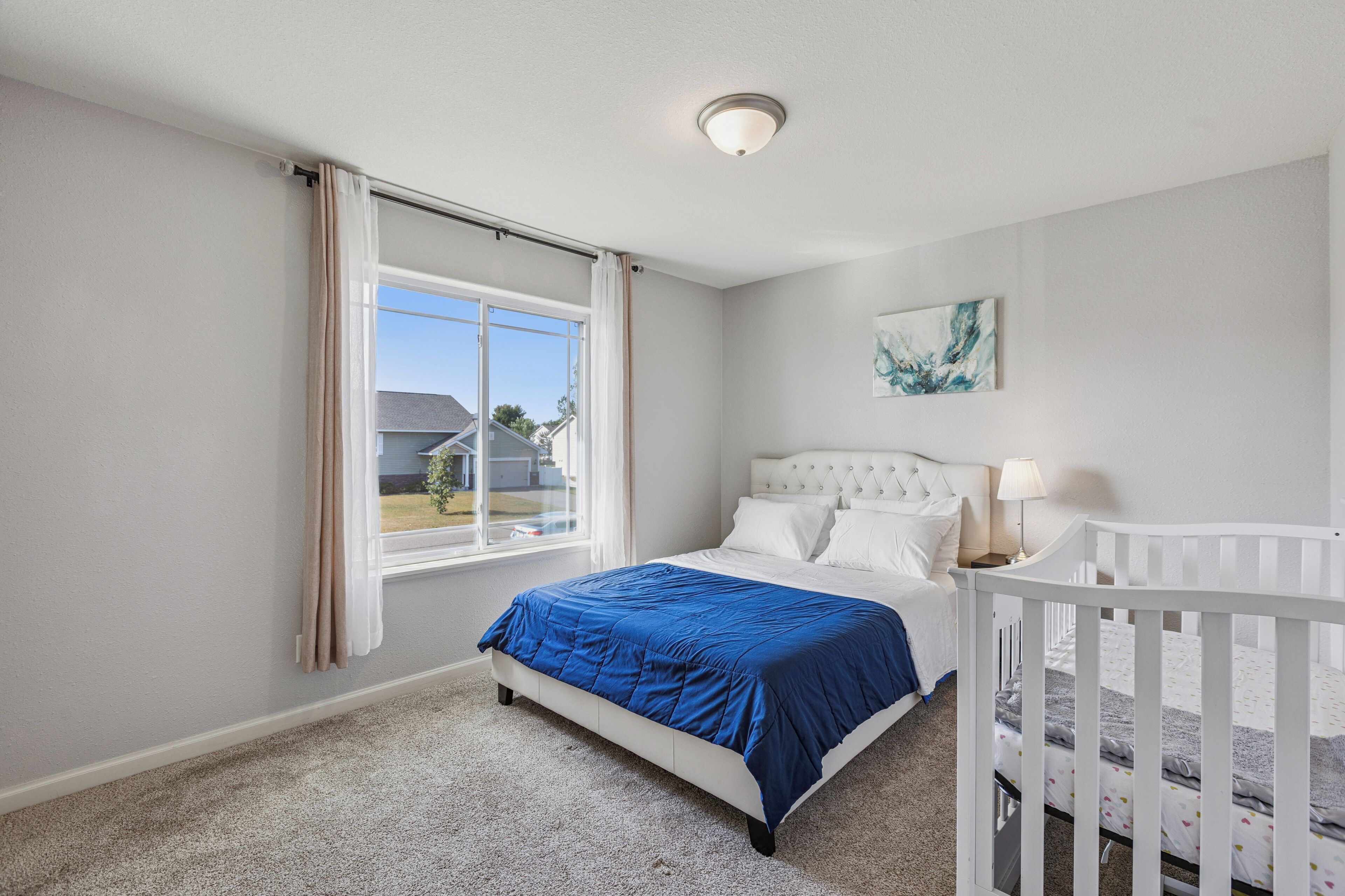 A bedroom with a large window, a bed with a blue bedspread, a crib, and a white headboard.