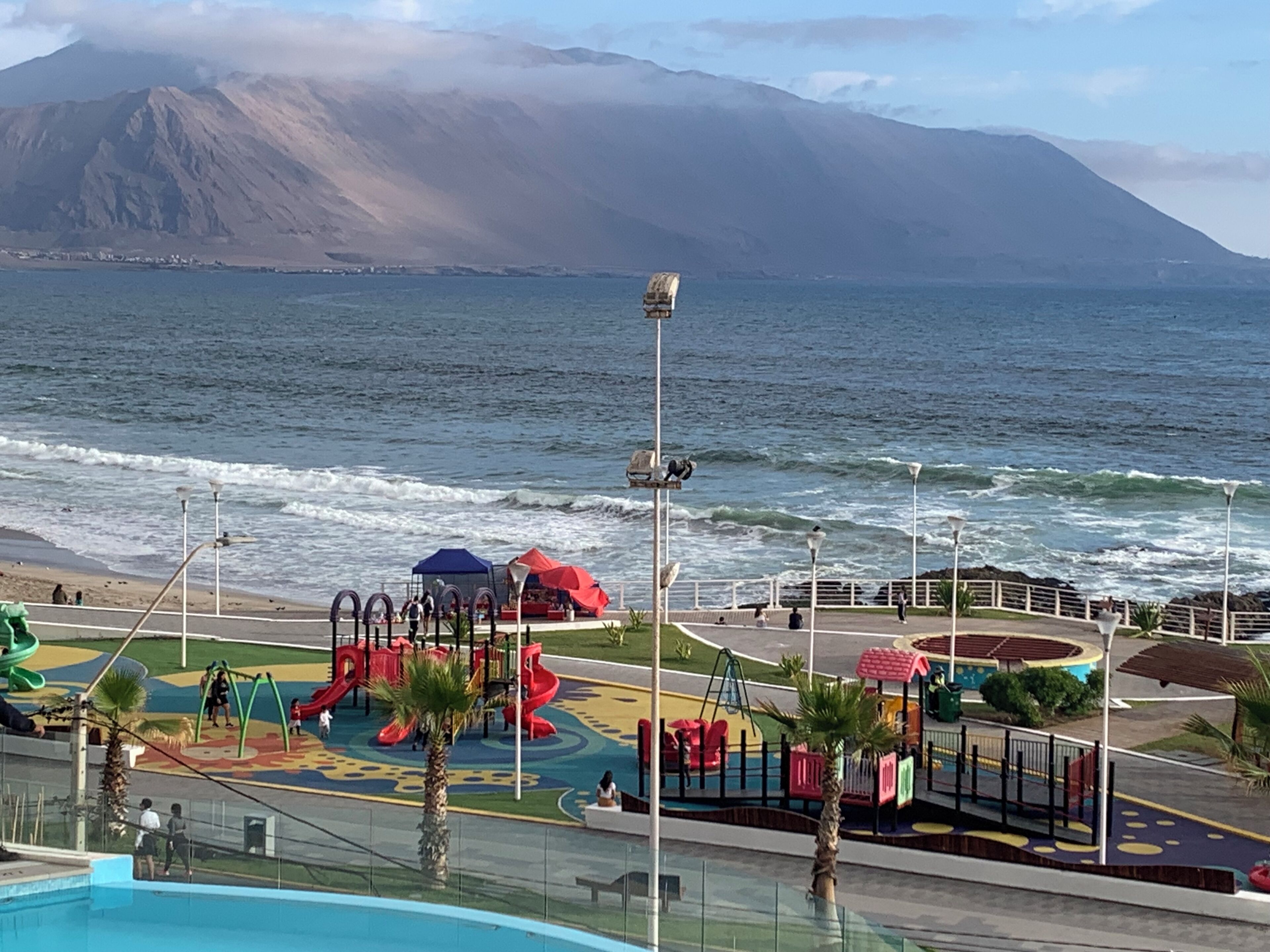 A coastal playground with slides and swings, a pool, and a view of the ocean and mountains.