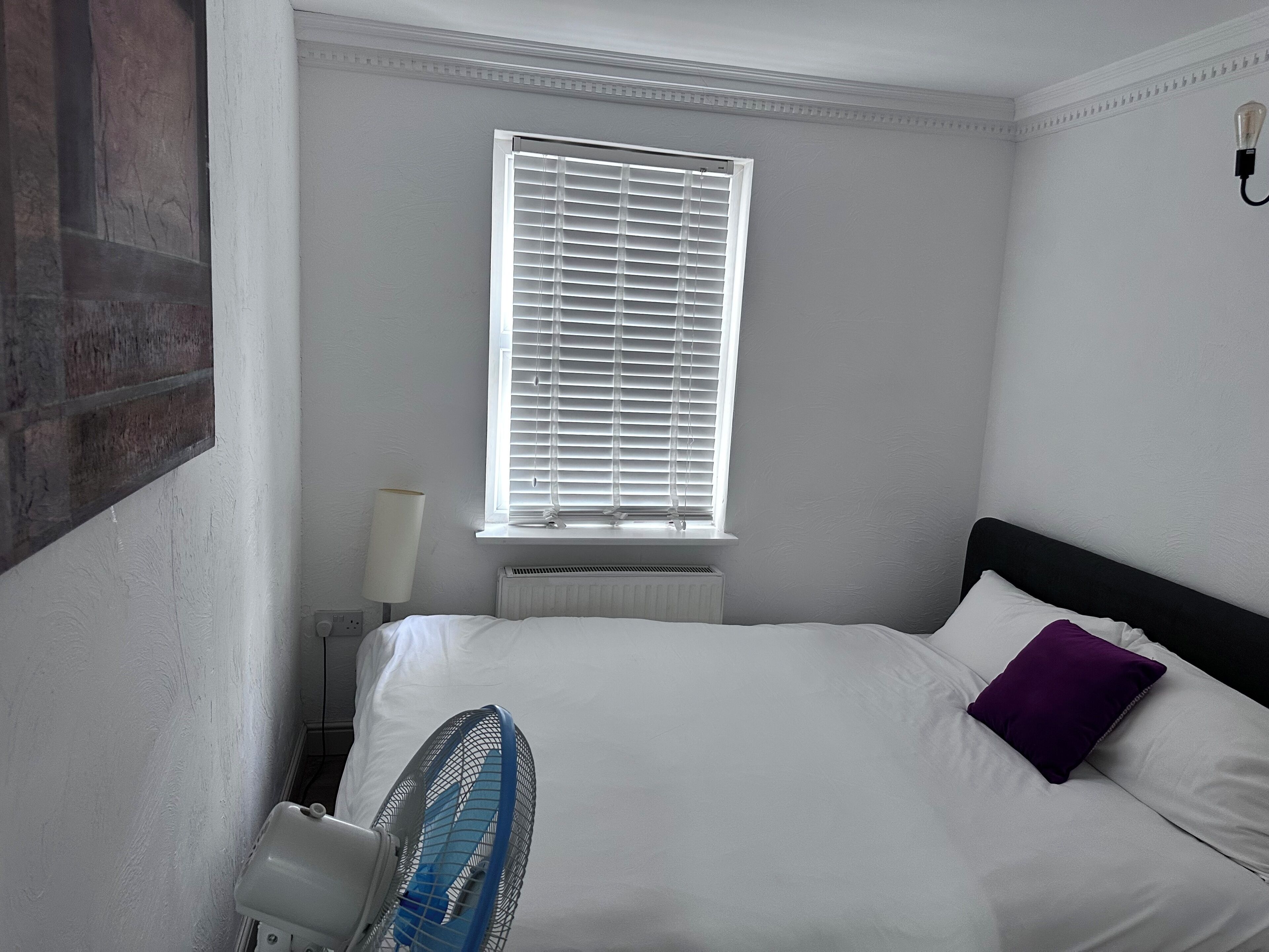 A hotel room with a bed, a window with blinds, a radiator, a lamp, and a wall-mounted light fixture.
