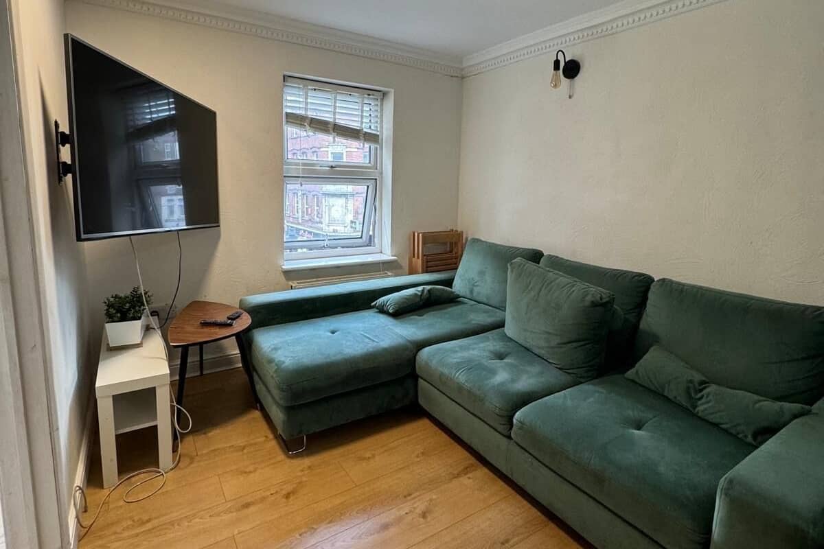 A living room with a green sofa, a small side table, a flat-screen TV mounted on the wall, and a window with blinds.