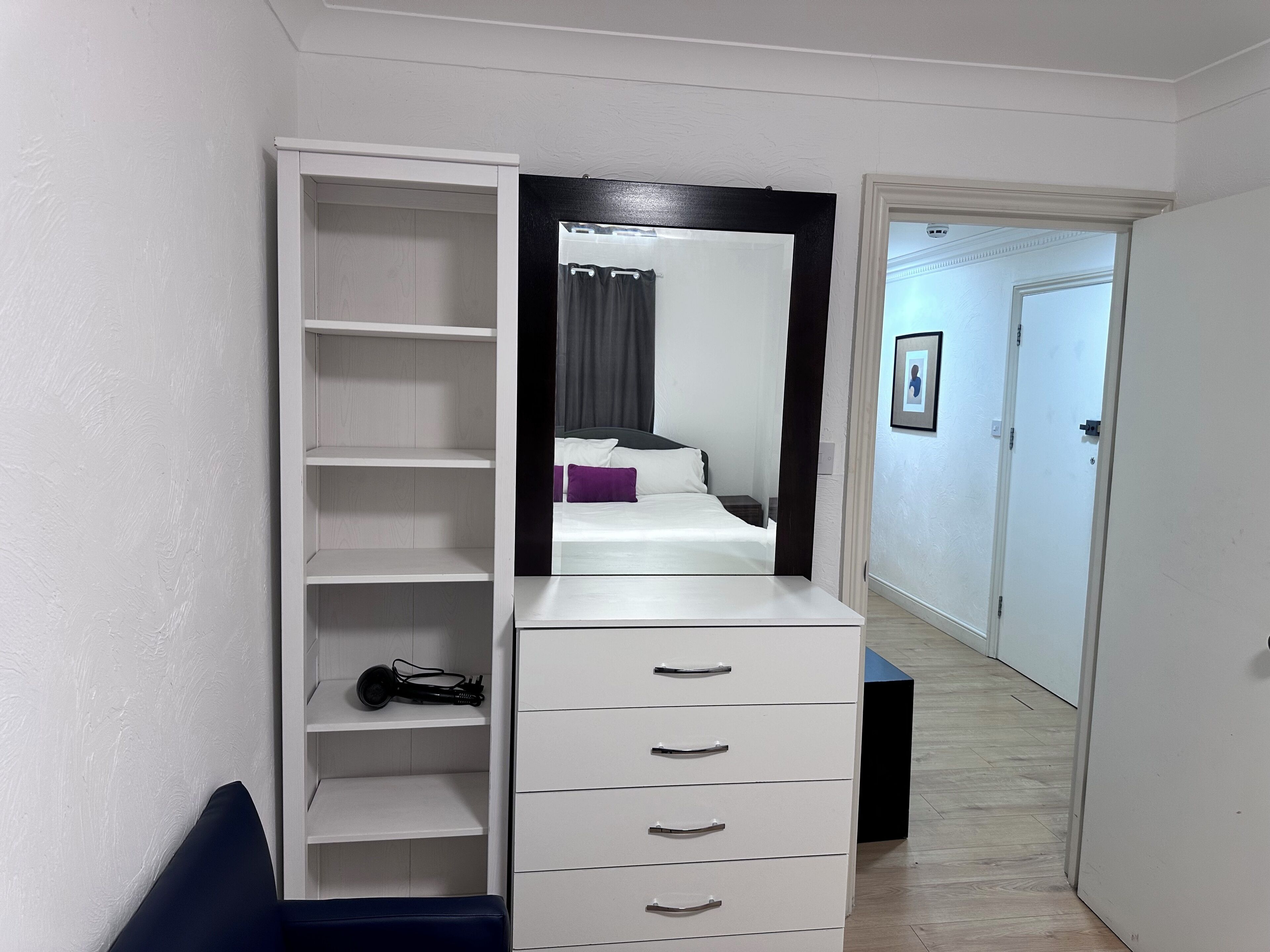 Un dormitorio con cama, un armario con varios cajones, un espejo y una puerta que da a otra habitación.