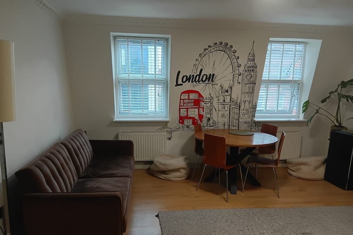 A living room with a brown leather sofa, a round wooden dining table with six chairs, two windows with blinds, a radiator, and a wall mural of London landmarks.