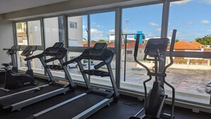 Un gimnasio con cintas de correr y elípticas, amplias ventanas y vistas a edificios y un estacionamiento.