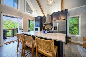 A modern kitchen with dark cabinets, a large island, and a view of the outdoors. - Couples Romantic Cabin! Hot Tub, Pizza Oven (Broken Bow)