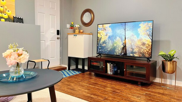 A living room with a wooden floor, a dining table with chairs, a TV on a stand, a cabinet, and a plant. - Private Basement + Self Check-in + Kids/Family + Parking (Bedford)