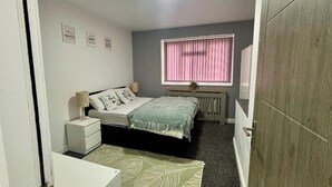 Een slaapkamer met een bed, nachtkastjes, een radiator, een raam met roze jaloezieën en een deur.