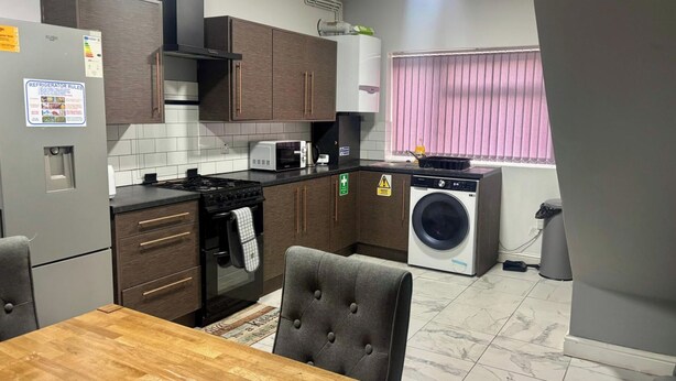 Een functionele keuken met een kookplaat, magnetron en wasmachine, met donkerhouten kastjes en een betegelde spatwand.