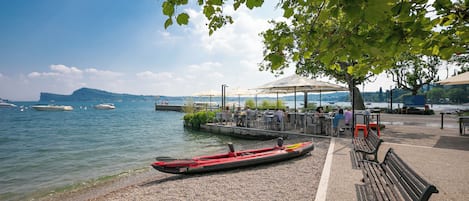 A lakeside park with a row of benches, a canoe, and a café area with tables and umbrellas.